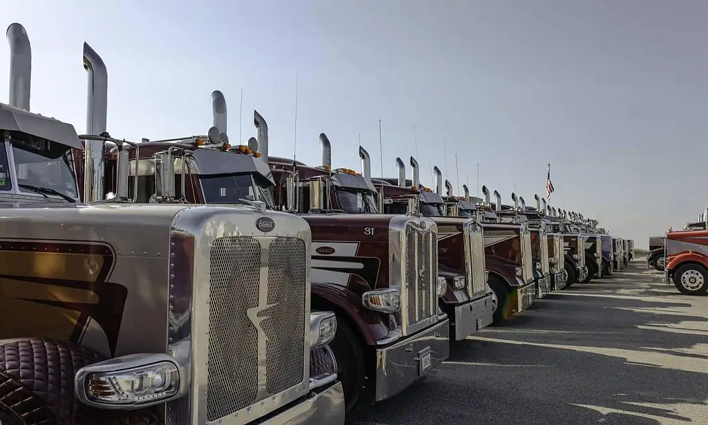 A long row of Farmers Oil Peterbilt tractors in maroon, chrome and cream lined up under a clear sky